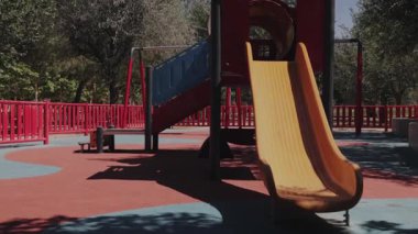 Canlı kırmızı ve sarı bir kaydırağın, bankların ve çocukların oynamadığı boş bir oyun parkının video görüntüleri. Gerçek zamanlı olarak kaydedildi, sükunet ve sessizlik sergileniyor..