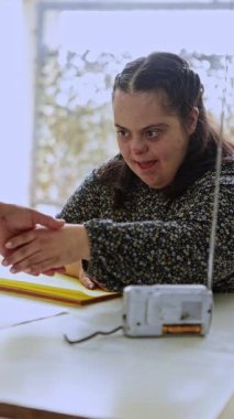 Down sendromlu genç bir kadın yaratıcı projesine odaklanıyor. Parlak, güneşli bir masada, elinde sanat malzemeleri ve bir teyp ile oturuyor..