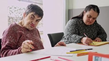 Zihinsel engelli iki kadın vatandaş, zihinsel gelişim faaliyetlerine katkı ve konsantrasyon göstererek, zihinsel sağlık merkezindeki bir masada çizim ve renklendirme faaliyetlerine odaklanmıştır.