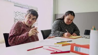 Zihinsel engelli iki kadın vatandaş, zihinsel gelişim faaliyetlerine katkı ve konsantrasyon göstererek, zihinsel sağlık merkezindeki bir masada çizim ve renklendirme faaliyetlerine odaklanmıştır.