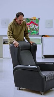 Akıl hastanesinde çalışan yetişkin bir erkek çocuk arka planda dekor gibi bir çocuğun olduğu bir odada yüksek koltuğu hareket ettiriyor. İfadesi düşünceli ve odaklanmış, bir anlık derin düşüncelere dalmış.