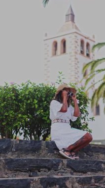 Şapkalı neşeli orta yaşlı bir kadın, İspanya 'nın Kanarya Adaları, Fuerteventura bölgesindeki tarihi bölgede güneşli bir günün tadını çıkarıyor. Yeşillik ve kilise kulesi fevkalade bir zemin oluşturuyor..