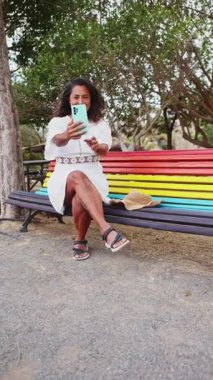 Neşeli orta yaşlı kadın, Fuerteventura 'da canlı, renkli bir bankta otururken akıllı telefonuyla selfie çekmekten hoşlanıyor..