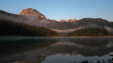 Karadağ 'ın dingin dağ manzarasının büyüleyici hava manzarası, açık mavi gökyüzünün altında sisli ormanlar ve engebeli tepelerle çevrili pitoresk bir göle ev sahipliği yapıyor.
