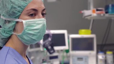 Cerrahi maske ve şapka takan bir hemşire modern bir hastane ameliyathanesine bakıyor. Arka planda tıbbi ekipman ve monitörler görülebiliyor ve sağlık hizmetleri ortamı vurgulanıyor.