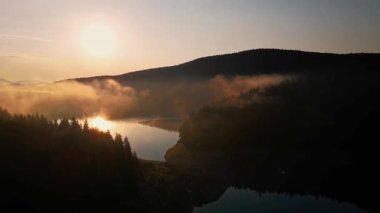 Karadağ 'daki ormanlı dağların üzerinden yükselen altın güneş, sakin bir göl üzerinde sürüklenen sabah sisinin içinden ılık bir ışık saçıyor ve büyülü ve sakin bir doğal atmosfer yaratıyor.