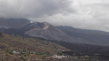 İspanya, La Palma 'da karanlık yamaçları ve bulutlu gökyüzü olan dramatik volkanik manzara. Ön planda verimli bitki örtüsü ve küçük binalar yer alıyor..