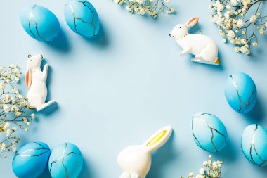 Paskalya yumurtalarının mermer taş efektli düz kompozisyonu, bahar çiçekleri, pastel mavi arka planda tavşanlar.