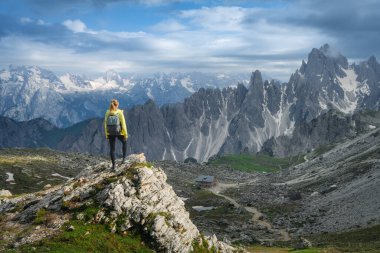 Sırt çantalı genç bir kadın dağ vadisinde yüksek dağlık tepeleri ve yazın günbatımında bulutlu gökyüzü olan bir kayanın üzerinde duruyor. Dolomitler, İtalya 'da yürüyüş. Seyahat, baharda seyahat.