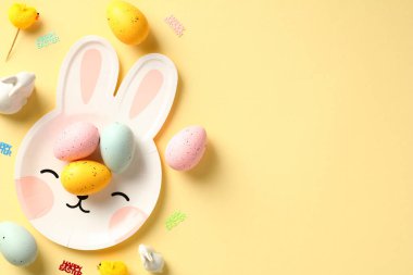 Renkli Paskalya yumurtalarının sarı arka planda tavşan şeklinde şirin tabakta olduğu düz bir kompozisyon. Mutlu Paskalya tebrik kartı tasarımı.