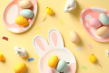 Mutlu Paskalyalar. Sarı arka planda paskalya yumurtası ve sofra takımı servis ediliyor. Düz yatış, üst görünüm.