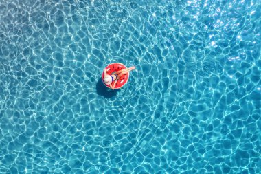 Yazın gündoğumunda mavi denizde kırmızı yüzme halkasıyla yüzen bir kadının hava görüntüsü. Kızlı tropik manzara, gök mavisi suyla okyanus, kumlu sahil. Üst Manzara. İtalya 'nın Sardunya adasında tatil
