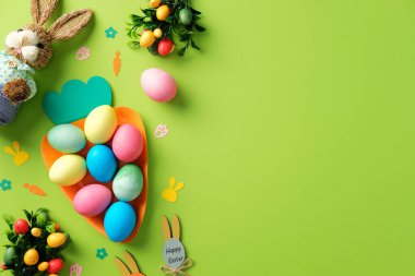 Mutlu Paskalyalar. Havuç şeklindeki tepside renkli Paskalya yumurtaları ve yeşil arka planda süslü püslü oyuncaklar..