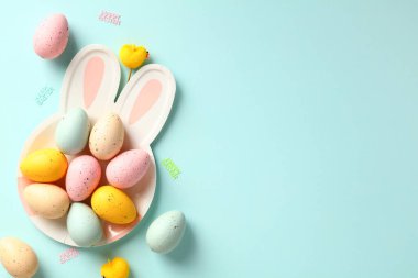 Tabakta renkli Paskalya yumurtaları ve pastel mavi arka planda tavşan kulakları. Düz konum, üst görünüm, kopyalama alanı.