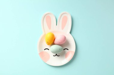 Pastel mavi arka planda renkli Paskalya yumurtaları olan tavşan şeklinde bir tabak. Düz yatış, üst görünüm.
