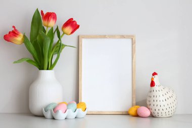 Paskalya evi dekorasyonları masada. Resim çerçevesi maketi, dekoratif tavuk, Paskalya yumurtaları, beyaz duvar arka planında lale vazosu..