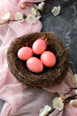 Kuş yuvasına yerleştirilmiş pembe Paskalya yumurtaları. Taze kiraz çiçekleri ve dokulu taş arka planda pembe bir kumaş..