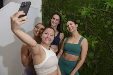 Spor giyim sektöründe dört mutlu kadın yeşil duvarlı bir spor salonunda pilates dersinden sonra akıllı telefonuyla selfie çekiyor.