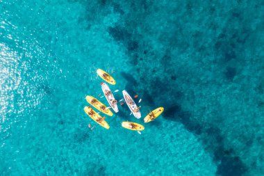 Aerial view of yellow kayaks in blue sea at sunset in summer. People on floating canoes in clear azure water. Sardinia island, Italy. Tropical landscape. Sup boards. Active travel. Top view from drone