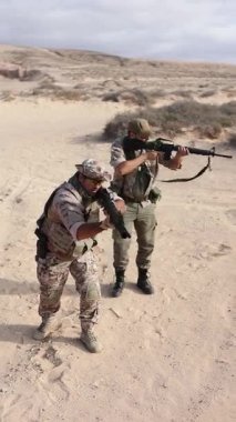 Kamuflaj üniforması giymiş bir çift erkek asker aktif bir eğitim tatbikatında, silahlarını çöl ortamına doğrultuyor, hazırlık ve takım çalışması sergiliyorlar..