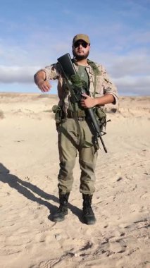 Kamuflaj kıyafetli bir erkek asker, açık bir gökyüzünün altındaki çöl ortamında elinde bir tüfekle selam veriyor..
