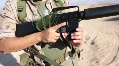 Tanınmayan bir erkek asker, kumlu bir çöl ortamında tüfeğine mühimmat yüklüyor ve görevine odaklanıyor..