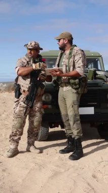 Biri tablet tutan iki asker, modern askeri operasyonlar ve teknolojik entegrasyonlar sergileyen çöl ortamındaki bir aracın yanında strateji tartışıyorlar..