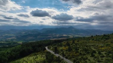İtalya 'daki Marche bölgesinin manzarası, yemyeşil tepeler, sık ormanlar arasında dolambaçlı bir yol ve parlak bir gökyüzü altında dramatik bulutlar. Seyahat ve doğa meraklıları için mükemmel.