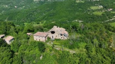 İtalya 'nın Marche bölgesinin yemyeşil tepelerinde yuva yapmış terk edilmiş bir çiftlik evinin havadan görünüşü. Canlı yeşillik ve geniş manzara huzurlu bir kırsal alan yaratır..
