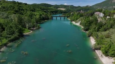 İtalya 'nın görkemli Marche bölgesinde yemyeşil ve dalgalı tepelerle çevrili turkuaz bir nehri kapsayan bir köprüyü barındıran sakin bir manzara.