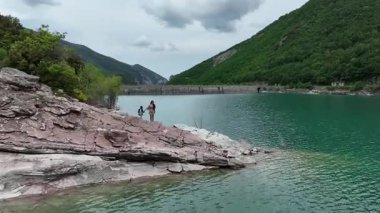 İtalya 'nın Marche kentinin havadan çekilmiş bir videosunda, bir anne ve çocuğu, kıvrımlı bir barajın yakınındaki turkuaz bir gölün kenarında, yemyeşil tepeler ve dramatik dağ manzaralarıyla çevrili.
