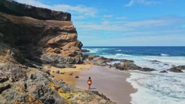 İspanya, Fuerteventura 'daki Tindaya Sahili' nin kumlu ve kayalık kıyılarında, Playa Tebeto olarak da bilinen kadının arka görüntüsü. El değmemiş sahil şeridi canlı mavi bir okyanusa karşı engebeli uçurumlara sahiptir..