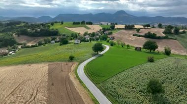 İtalya 'nın Marş bölgesinin kayan kırsal manzarasında yemyeşil tarlalar, dolambaçlı bir yol ve bulutlu bir gökyüzünün altındaki uzak dağlar sergileniyor.