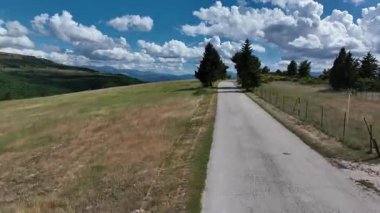 İtalya 'nın Marche bölgesinde pitoresk bir manzara. Video, yemyeşil tepeler, dağınık ağaçlar ve kabarık bulutlarla dolu canlı mavi bir gökyüzünün altında dolambaçlı bir yol yakalıyor.