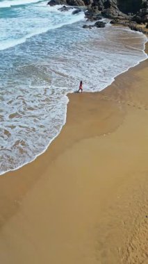 Kanarya Adaları, Fuerteventura, İspanya 'daki Tindaya Sahili' nin kumlu kayalık sahillerinde dalgalı denizde yürüyen bir kadının uzaktan kumandalı hava aracı görüntüsü. Kıyı şeridi dalgalı mavi bir okyanusa karşı engebeli uçurumlara sahiptir..