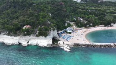 İtalya 'nın Marche bölgesindeki yemyeşil uçurumların altında yer alan ve canlı şemsiyeler ve berrak mavi suları gösteren pitoresk bir sahil manzarası.