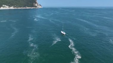 Bir yelkenli İtalya 'nın Marche bölgesinin açıklarında Adriyatik Denizi' nin turkuaz sularında yol alır. Bu manzarada yemyeşil tepeler ve kalabalık bir plaj var.