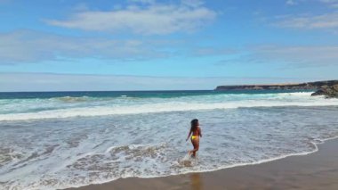 Renkli bikinili bir kadın tatillerde Fuerteventura, Kanarya Adaları 'ndaki Tindaya Sahili' nde mutlu bir şekilde koşup oynuyor. Kıyı şeridi dalgalı mavi bir okyanusa karşı engebeli uçurumlara sahiptir..