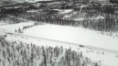 Hava aracı görüntülerinde, Norveç 'te kış boyunca yoğun bir İskandinav ormanında karla kaplı bir otoyol boyunca seyahat eden yalnız bir minibüs görüntüleniyor..