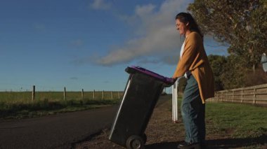 Günlük kıyafet giymiş bir kadın, kırsal bir ortamda çöp tenekesini itiyor. Güneş sıcacık bir parıltı saçar, yuvarlanan tarlalar ve açık bir gökyüzü arasında rutin bir görevi güçlendirir.