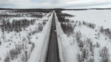 Hava aracı görüntüleri karlı, tenha bir İskandinav yolu boyunca bir araba yolculuğunu ve kenarına park edilmiş bir minibüsü yakalıyor. Geniş, buz gibi bir manzarayla çevrili.