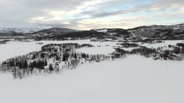 Norveç 'teki el değmemiş karlı bir arazinin insansız hava aracı görüntüsü, kar dolu ormanlar ve uzak dağlarla çevrili geleneksel İskandinav evlerinin görüntüsü..