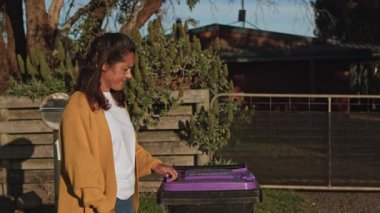 Günlük kıyafet giymiş bir kadın çöpü dışarı çıkarır. O, güneşli bir arka planda yer alan çöp kutusunun mor kapağını kaldırırken burnunu tutuyor.