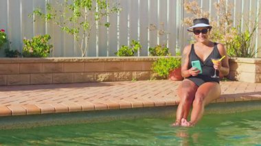 Bir kadın havuzun kenarında rahatça oturur, bacakları daldırılmış, ferahlatıcı bir kokteyl tutar ve akıllı telefonuna bakar. Güneşli bir günün tadını çıkarıyor, güneş gözlüğü ve şapka takıyor.