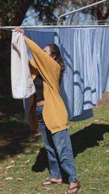 Gündelik giysiler içindeki bir kadın, sıcak güneşin altında çamaşır ipine asılır. Açık havada, yeşil ve doğal ışıkla çevrili günlük görevinin tadını çıkarır.