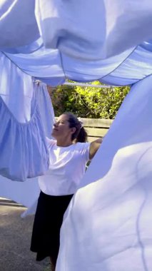 Bir kadın dışarıda çamaşır ipine yeni yıkanmış mavi çarşafları asarken gülümsüyor. Güneş ışığı çamaşırların arasından süzülüp günlük huzurlu bir rutin an yaratıyor.