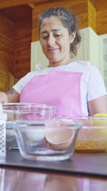 Pembe önlüklü bir kadın rahat, kırsal bir mutfakta kurabiye pişirmeye odaklanır. Çeşitli karıştırma kaseleri ve malzemeler tezgahta hazır, yemek pişirmenin zevkini yakalıyorlar.