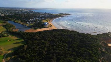Avustralya 'daki Büyük Okyanus Yolu boyunca uzanan ve geniş okyanus manzaraları, yemyeşil alanlar ve berrak bir gökyüzü altında sakin bir sahil kasabasının yer aldığı pitoresk bir sahil manzarası..