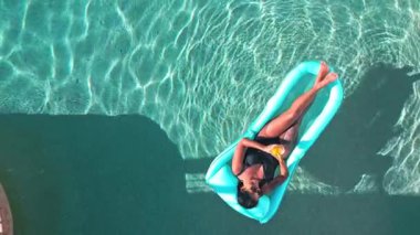 Bir kadın, sakin bir havuzda şişme bir şamandıranın üzerinde içkisini yudumlarken dinleniyor. Güneş ışığı suda hafif dalgalanmalar yaratır, sakin bir atmosfer yaratır, rahatlamak için idealdir.