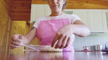 Pembe önlüklü bir kadın kurabiye hamurunu dikkatlice plastik bir ambalaja sarıp ahşap bir mutfağa koyuyor. Modern dekorlarla çevrili olarak işine odaklanmış görünüyor.